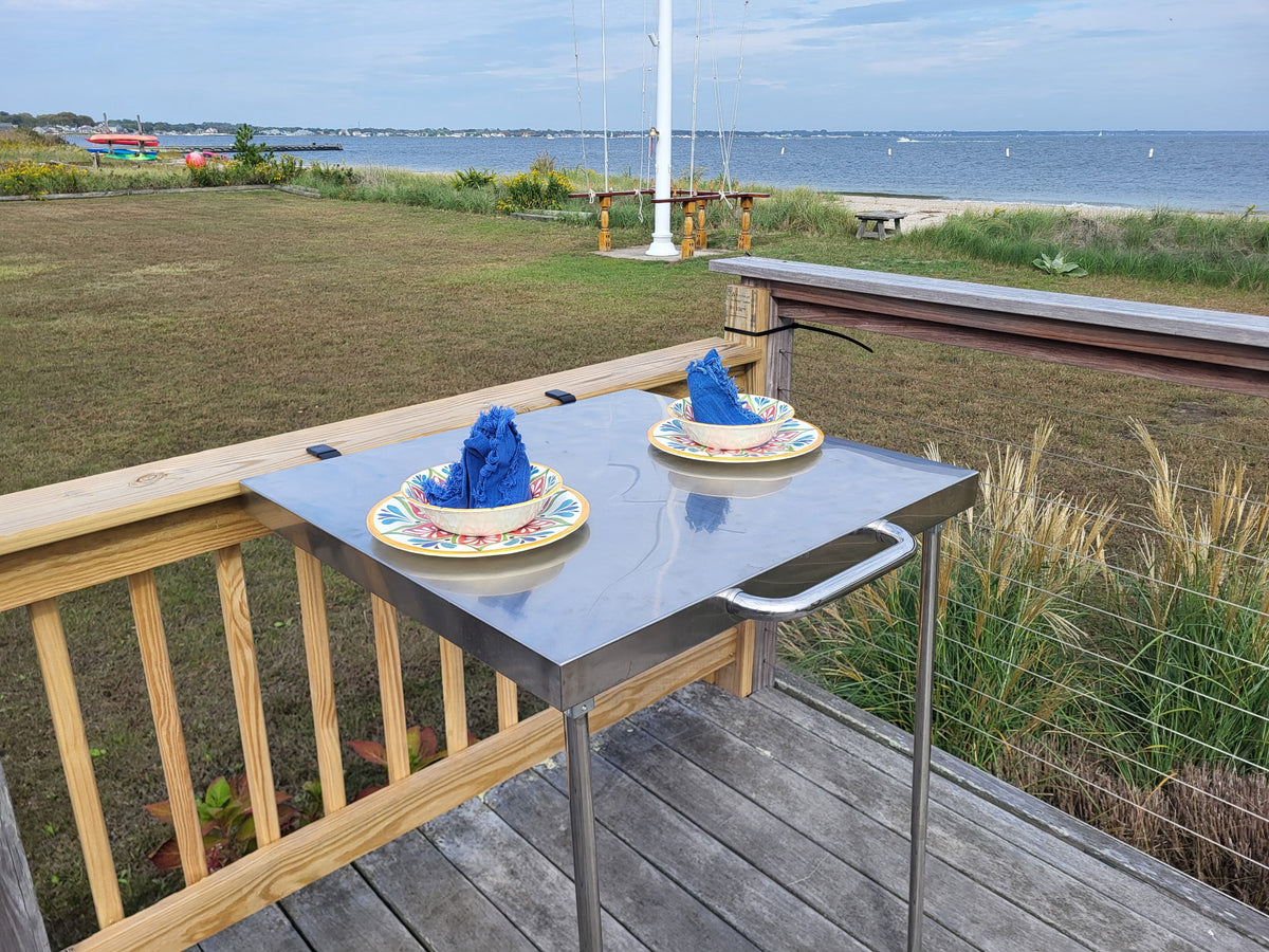 The Big John Table clamped to a waterfront deck railing, set with colorful plates and blue napkins, with the bay behind it
