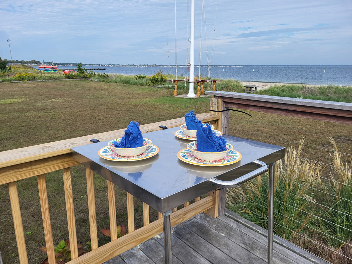 The Big John Table's stainless-steel top and handle, clamped to a wooden deck railing overlooking the water