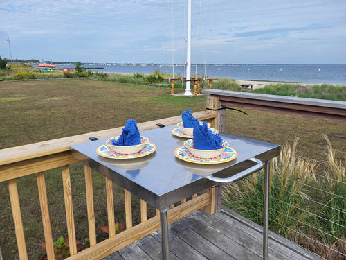 The Big John Table's stainless-steel top and handle, clamped to a wooden deck railing overlooking the water