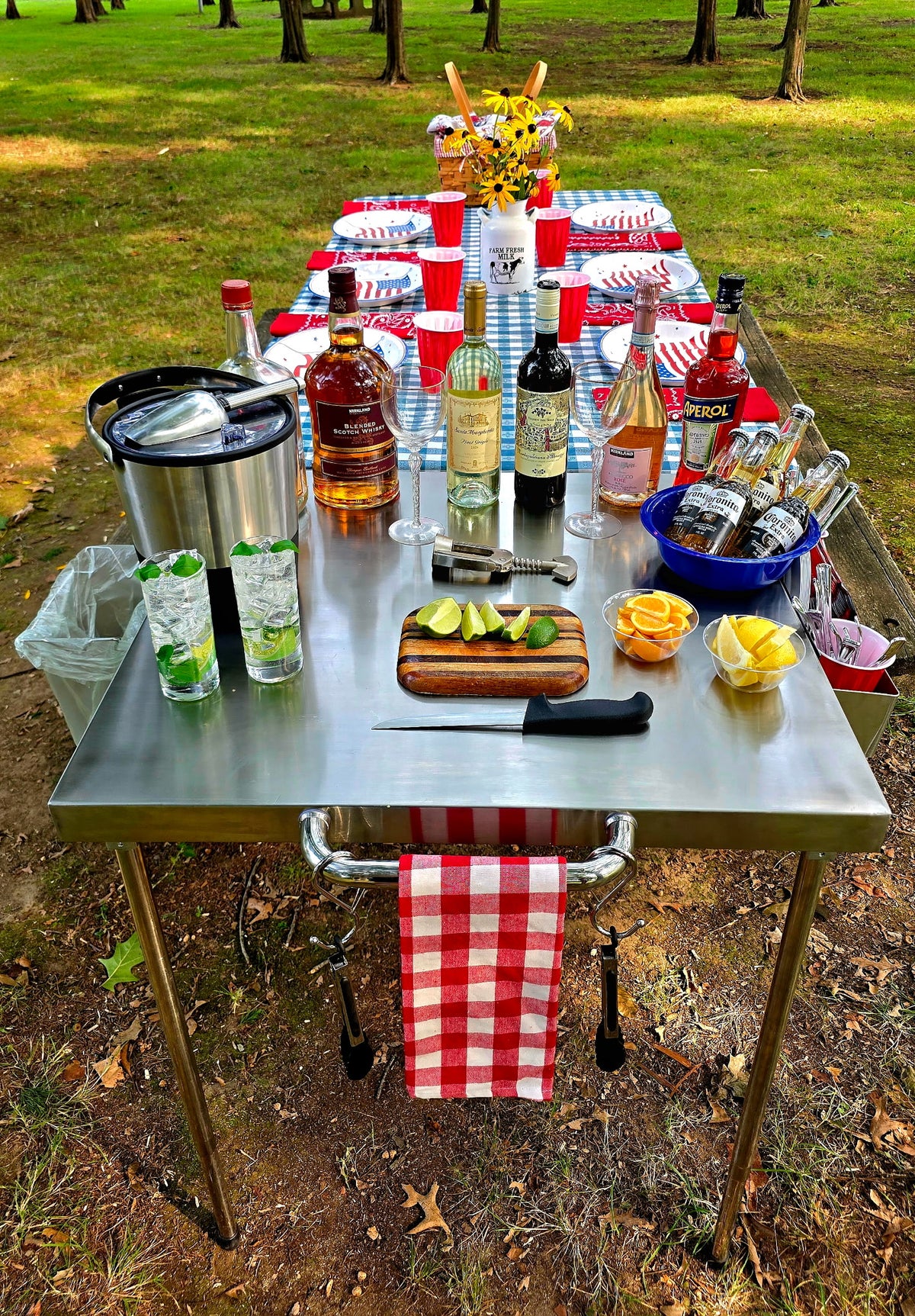 The Big John Table set up as an outdoor drink station with bottles, glasses, ice bucket and cut limes and lemons