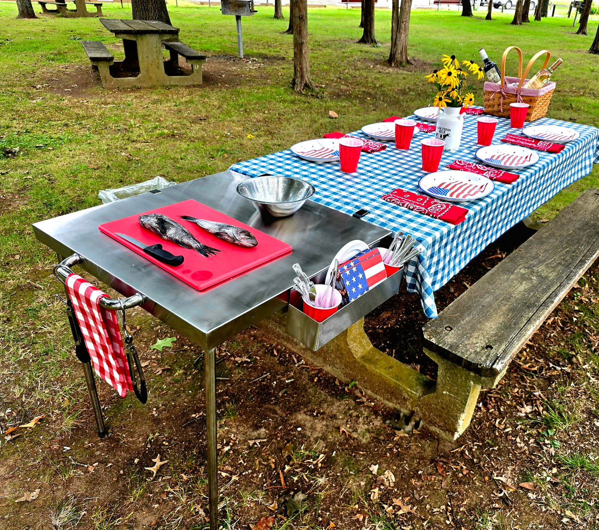 The Big John Table as a food prep station clamped to a picnic table set for a meal in the park