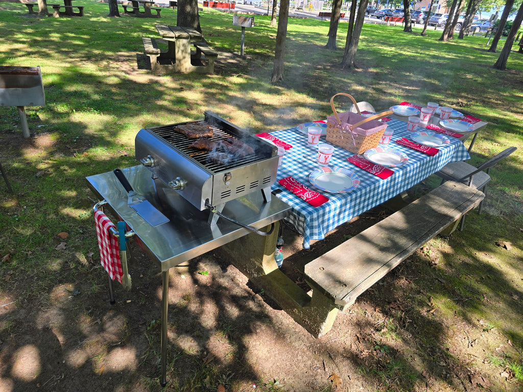 Portable gas grill cooking on The Big John Table attached to a picnic table set for a meal